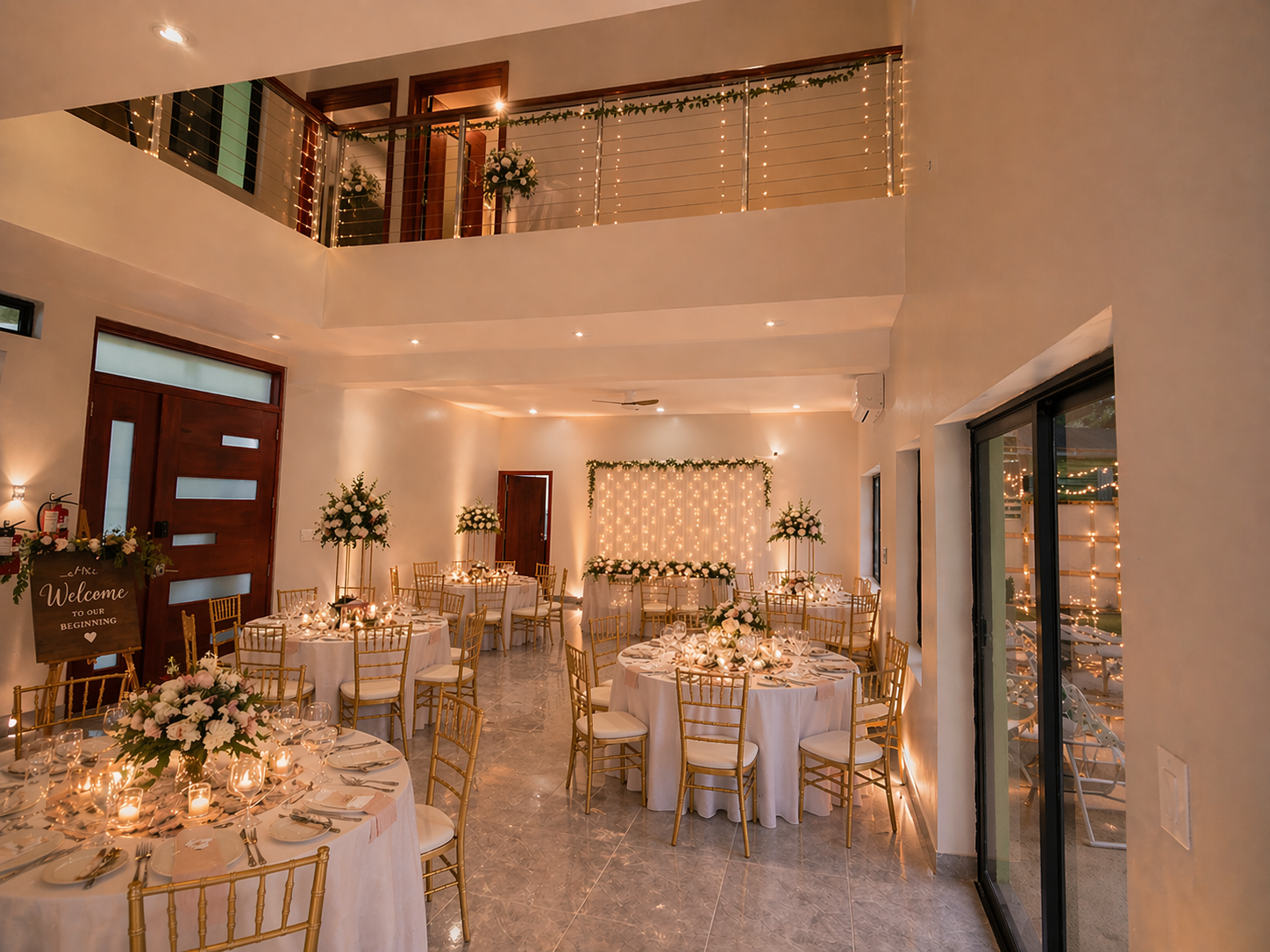 Private Event Venue in Placencia — MeMe's Place 8 MeMe's Place indoor event room set for a reception: round tables dressed in white linen, gold Chiavari chairs, tall centerpieces, fairy-light backdrop, welcome sign at entry.